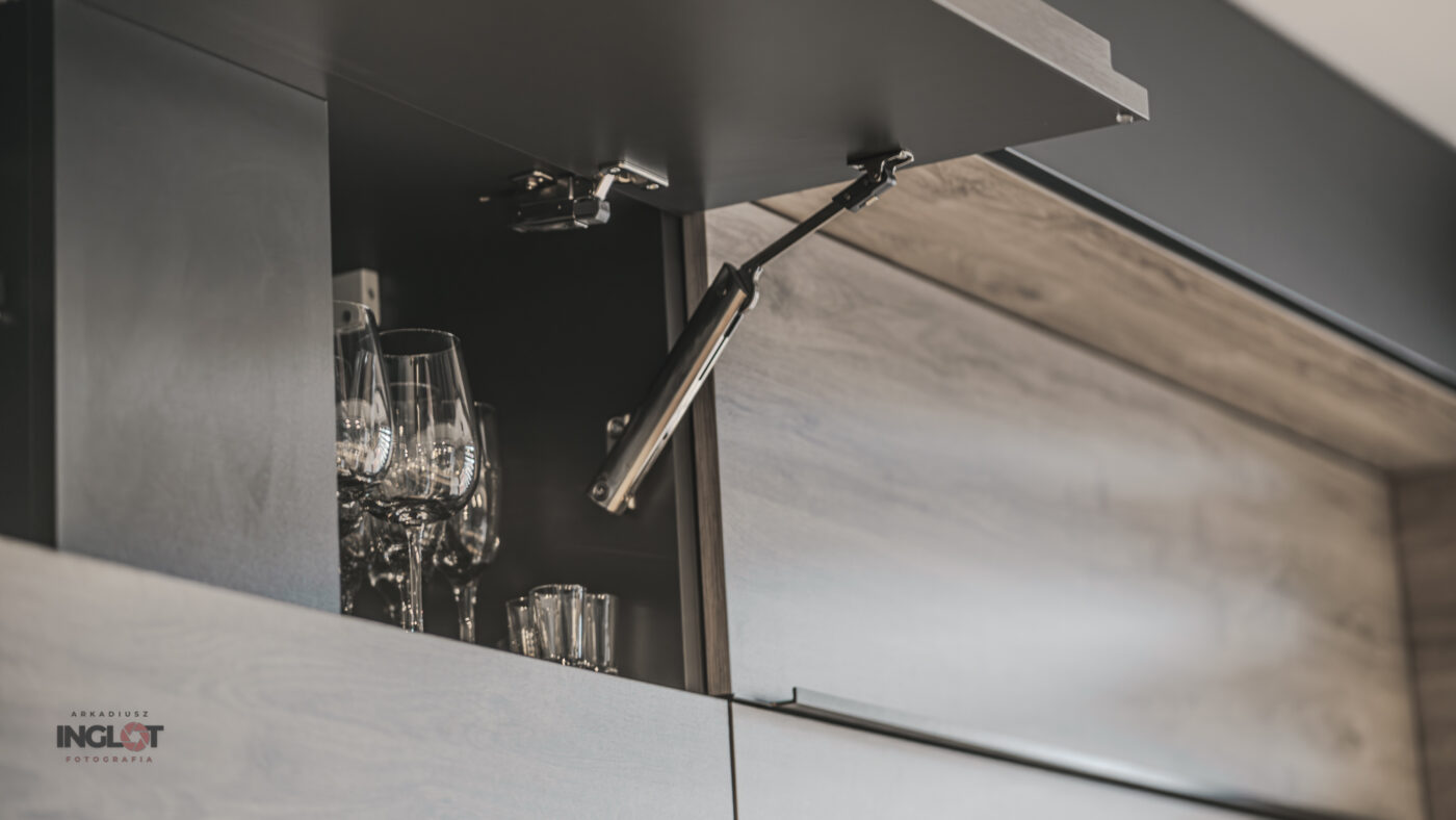 Zbliżenie na zawiasy w górnej szafce kuchennej z kieliszkami – gra światłem w fotografii wnętrz.