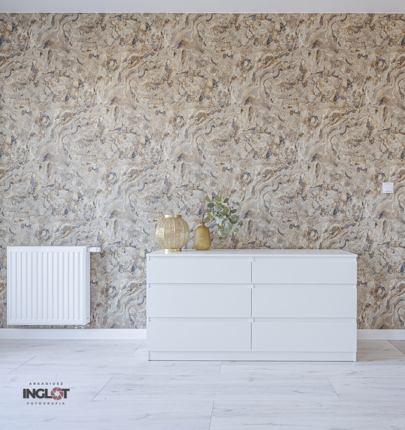 Stylowa biała szafka RTV ustawiona na tle oryginalnej, dekoracyjnej ściany. Minimalistyczny design mebla doskonale komponuje się z ciekawą fakturą lub wzorem ściany, tworząc elegancką i nowoczesną aranżację salonu.