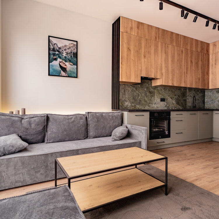 Boczne ujęcie salonu z szarą sofą, drewnianymi akcentami i aneksem kuchennym.