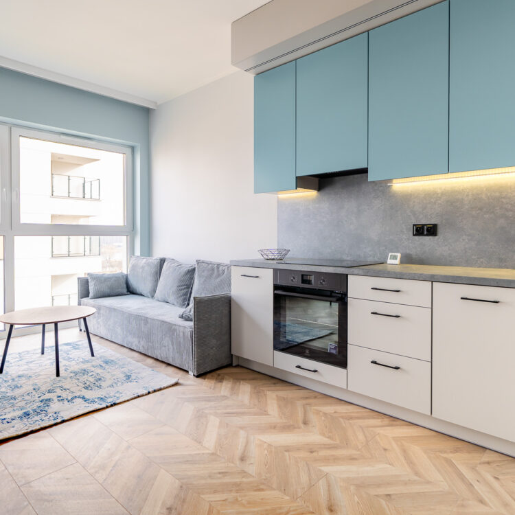Widok na kuchnię, strefę relaksu i wielkie okno, nowoczesna aranżacja wnętrza z dużą ilością naturalnego światła.
