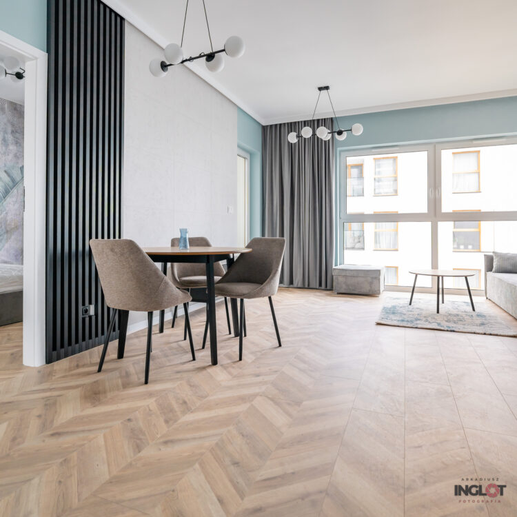 Widok na drewniany stół, strefę relaksu i wielkie okno, fotografia wnętrz podkreślająca naturalne światło i przestronność.